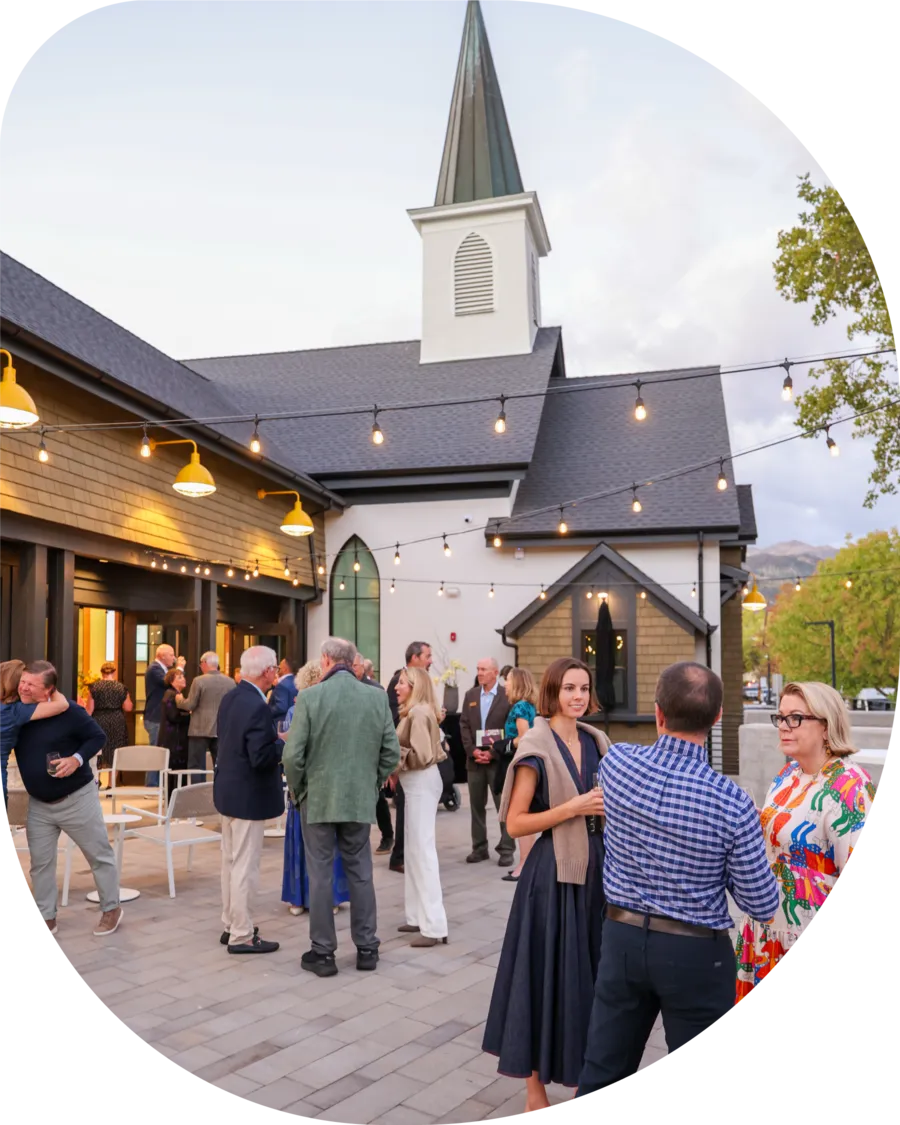 Outdoor gathering at a small church with people sitting and standing under stringed triangular flags on a sunny day.