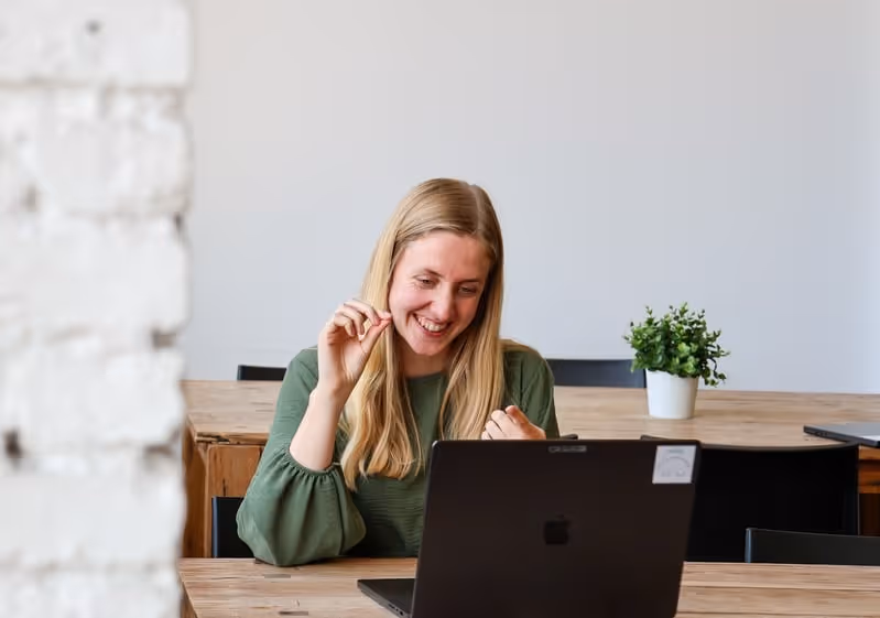 Coach die glimlachend een online gesprek voert achter haar laptop in een lichte werkruimte.