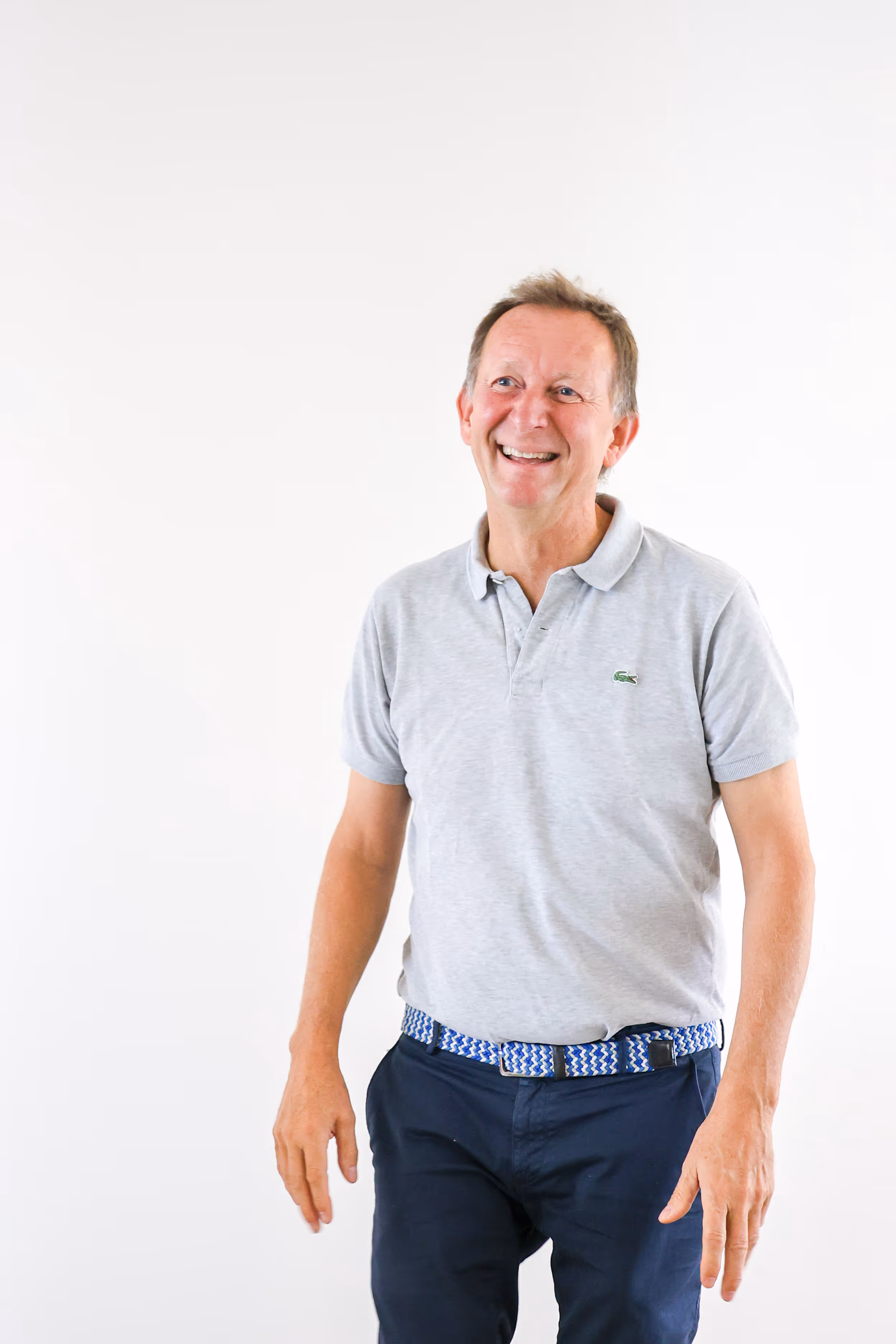 Man lacht, draagt lichtgrijze polo en donkere broek met blauwe riem, staande tegen witte achtergrond.
