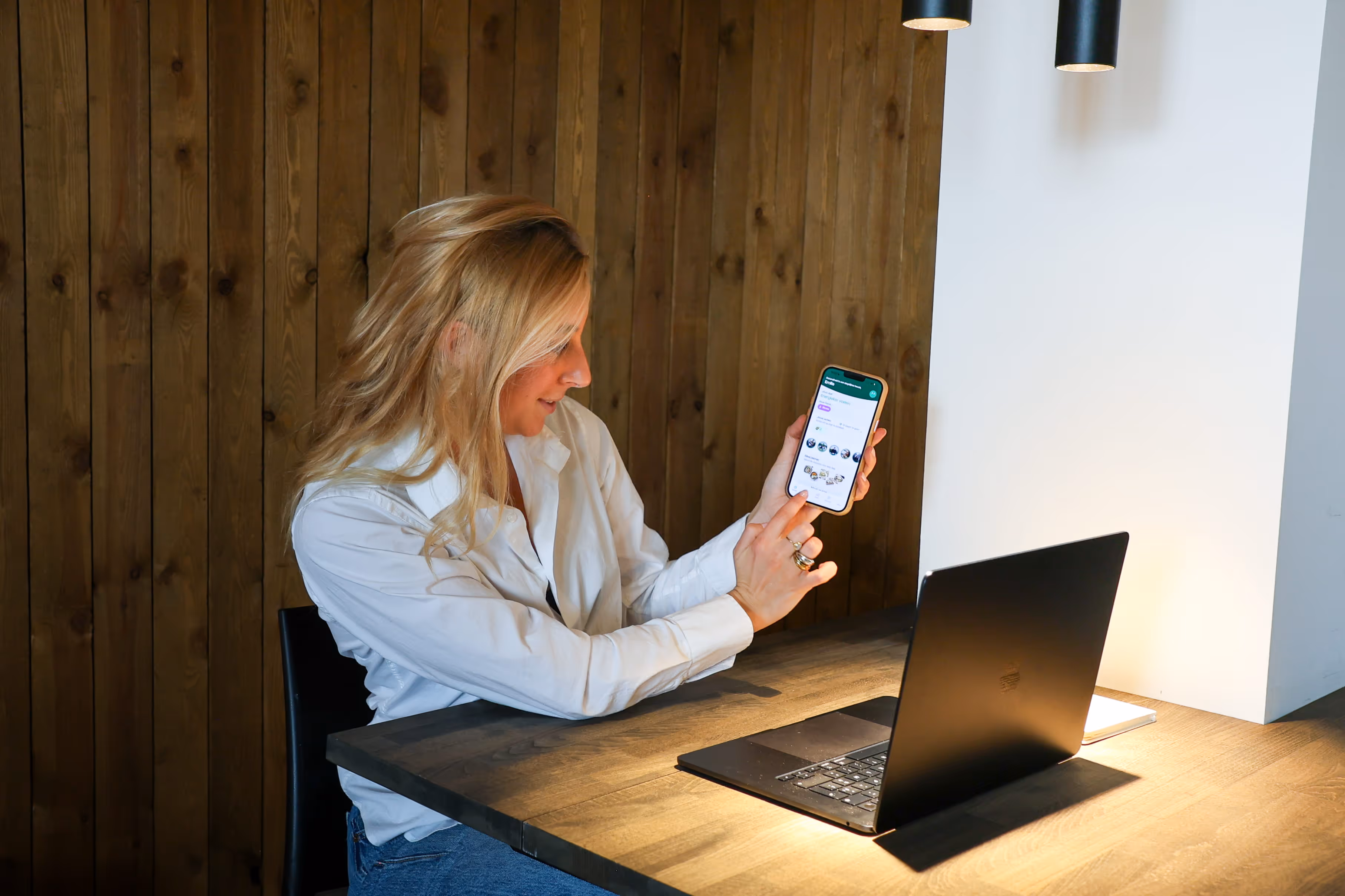Vrouw toont mobiel met Yoboo app en zit aan houten tafel met open laptop in een rustieke omgeving.