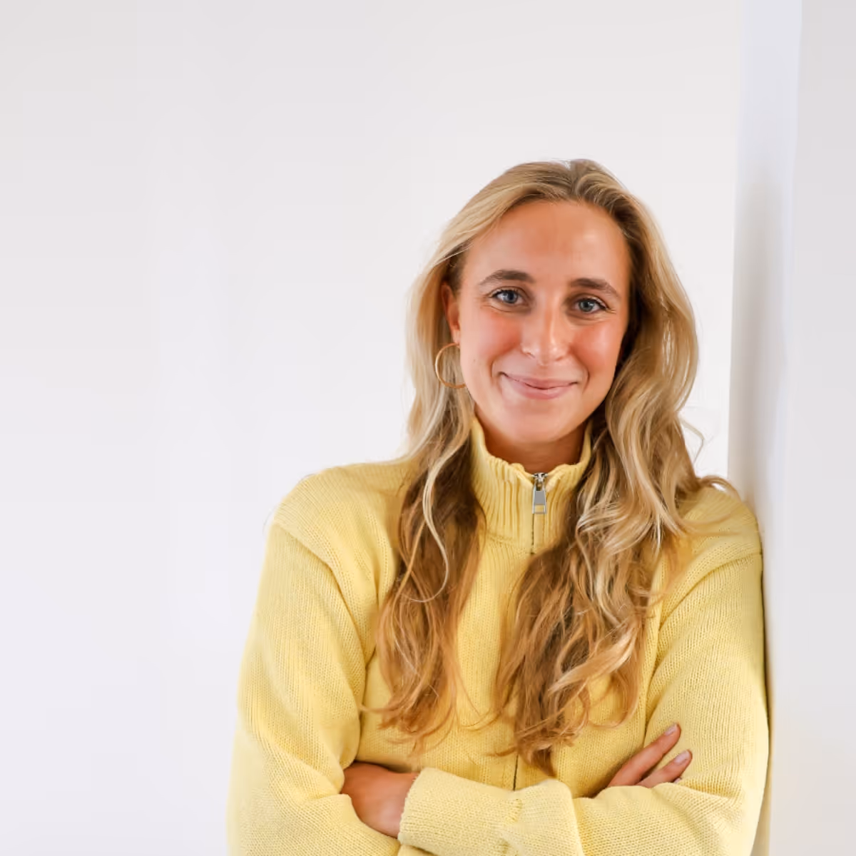 Vrouw met lang blond haar draagt gele trui en glimlacht met armen gekruist tegen een witte muur.