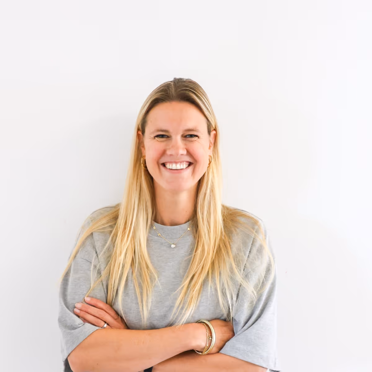 Vrouw met lang blond haar lacht, draagt grijze trui en heeft armen gekruist.