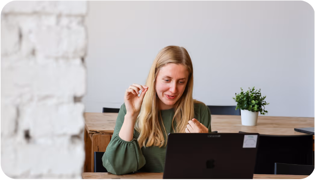 Vrouw met lang haar kijkt naar laptop en praat terwijl ze zit aan houten tafel.
