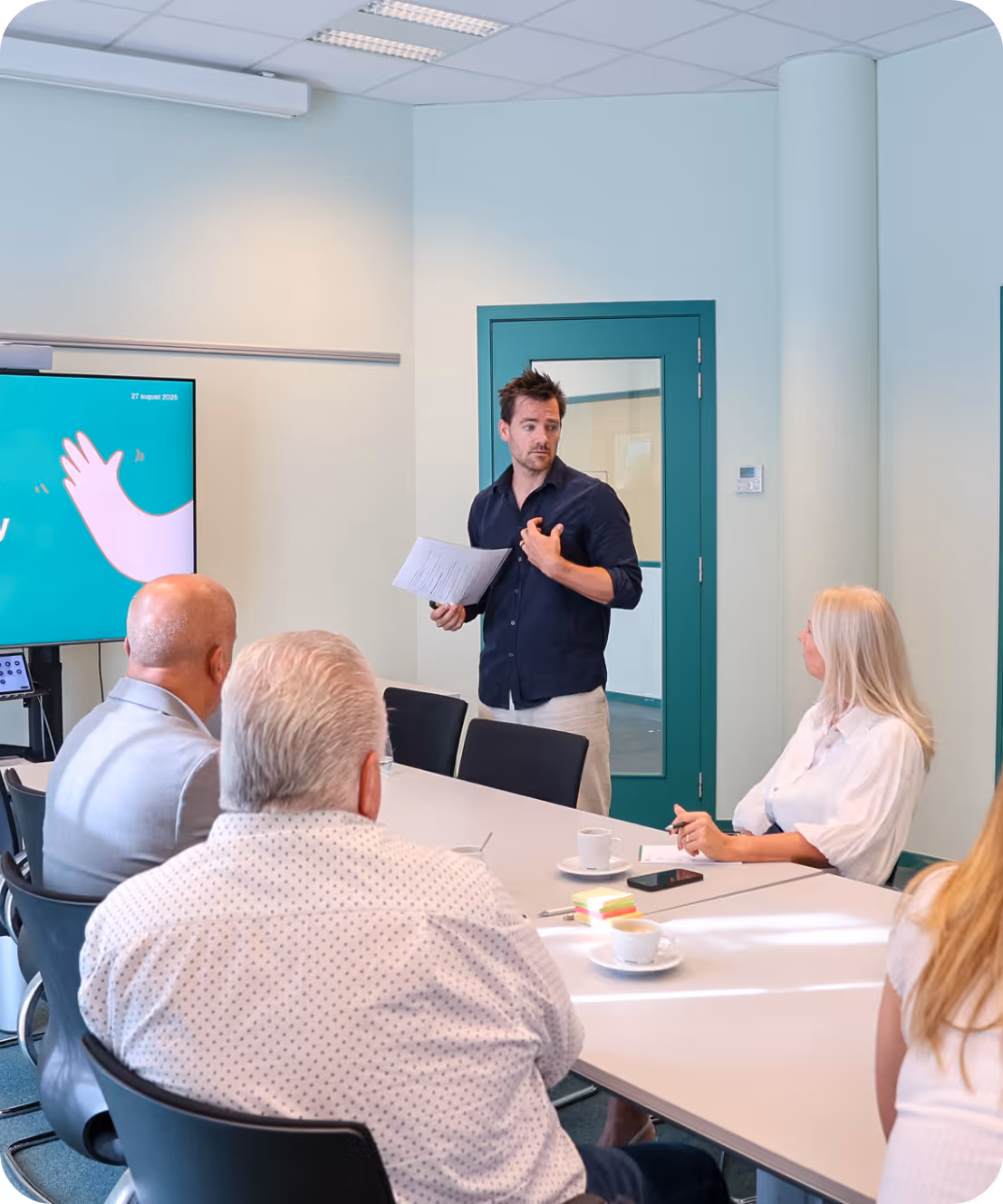 Man geeft presentatie aan vier collega’s rond vergadertafel in moderne kantoorruimte.
