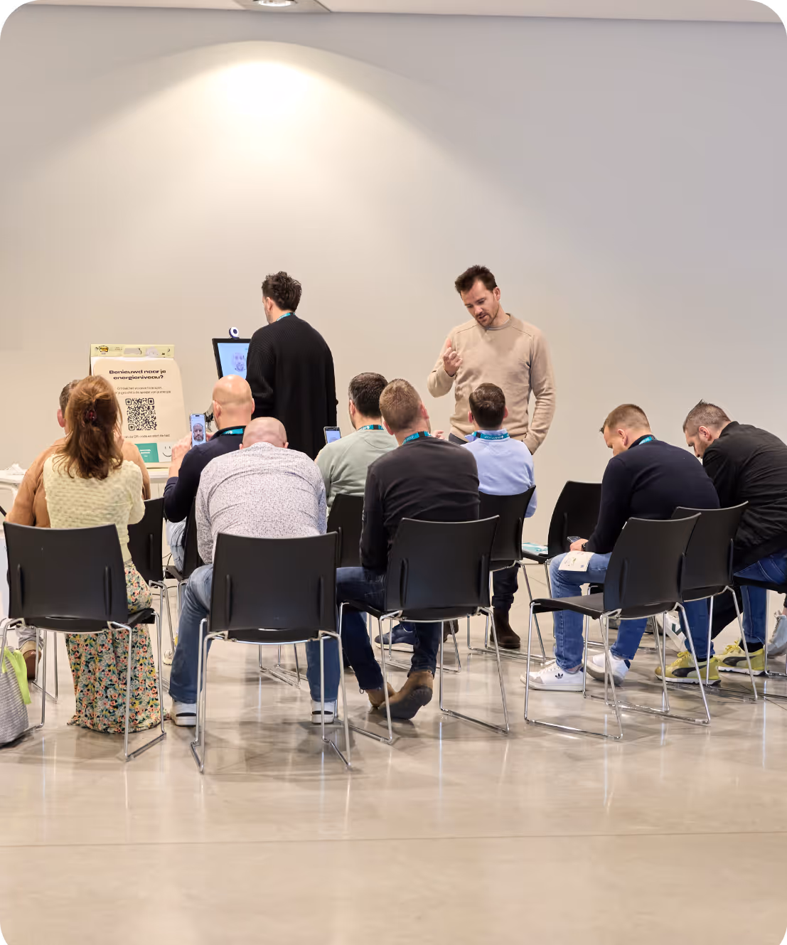 Groep mensen zit in stoelen, te luisteren naar twee mannen voor een informatiescherm in een zaal.