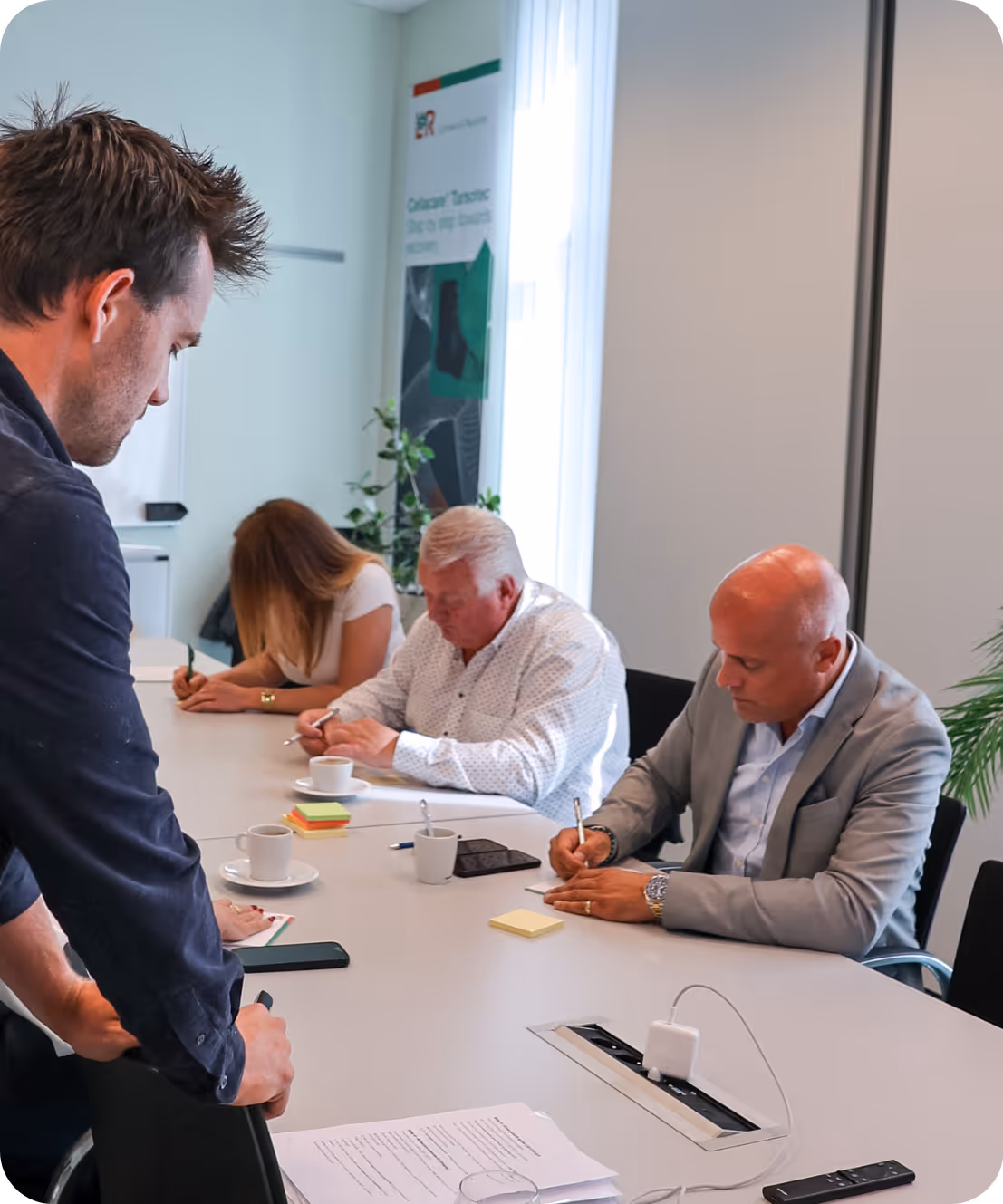 Vier mensen zitten aan vergadertafel en maken aantekeningen tijdens vergadering.