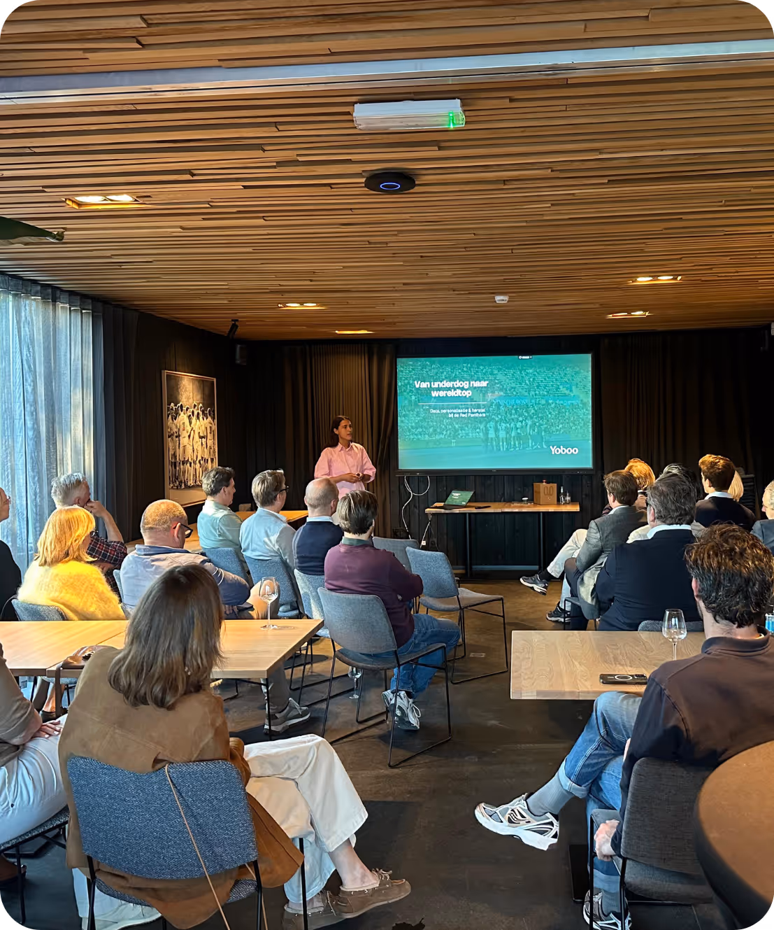 Presentator in roze overhemd geeft lezing aan zittend publiek in moderne conferentieruimte.