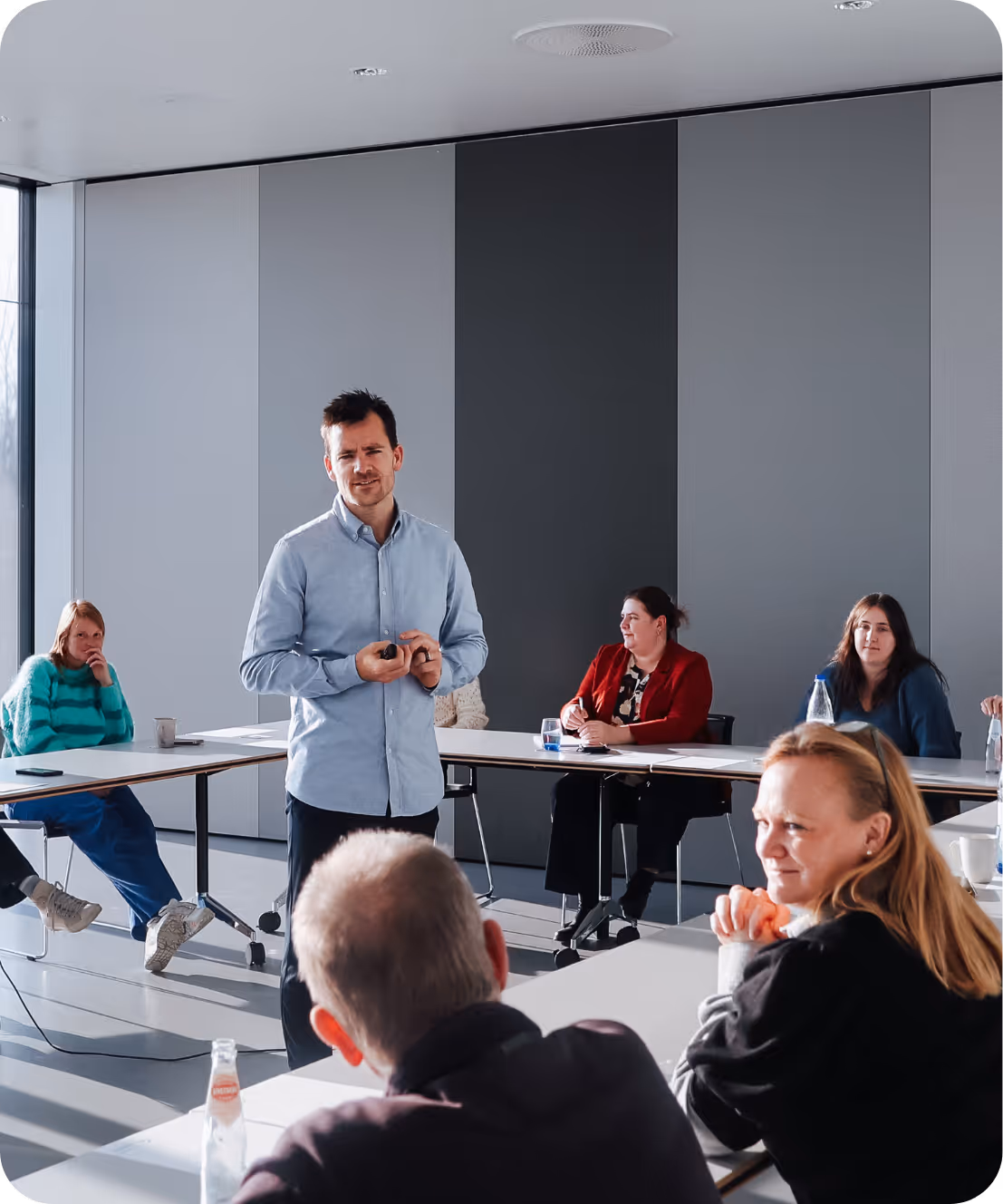 Man geeft presentatie aan een groep volwassenen die aan tafels in een moderne vergaderruimte zitten.