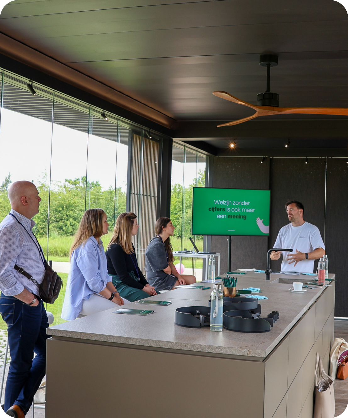 Groep mensen luistert naar een presentatie in een kamer met glazen wanden en groen uitzicht.