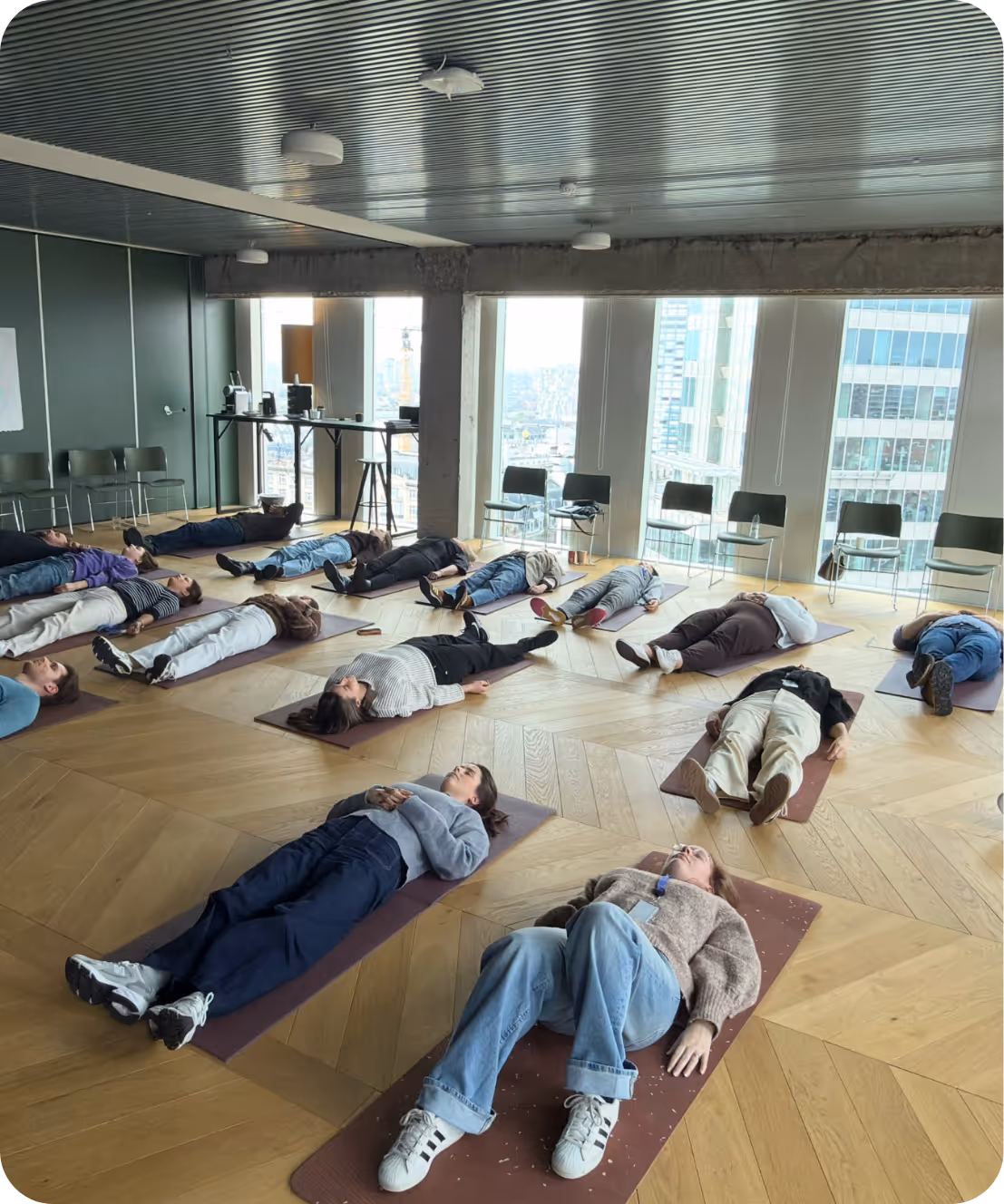 Een groep mensen ligt op yogamatten op een houten vloer in een lichte kamer met uitzicht op stadsgebouwen.