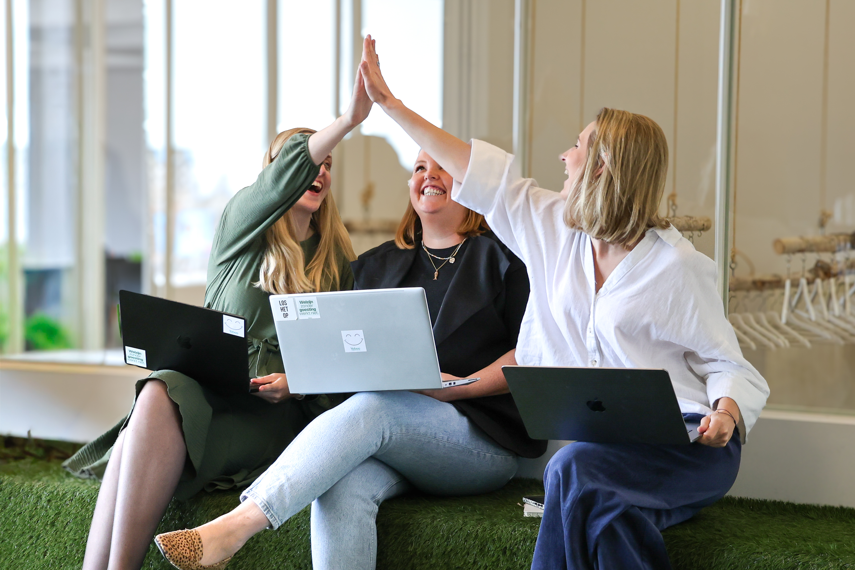 Drie vrouwen met laptops geven elkaar een high five terwijl ze op een groene bank zitten.