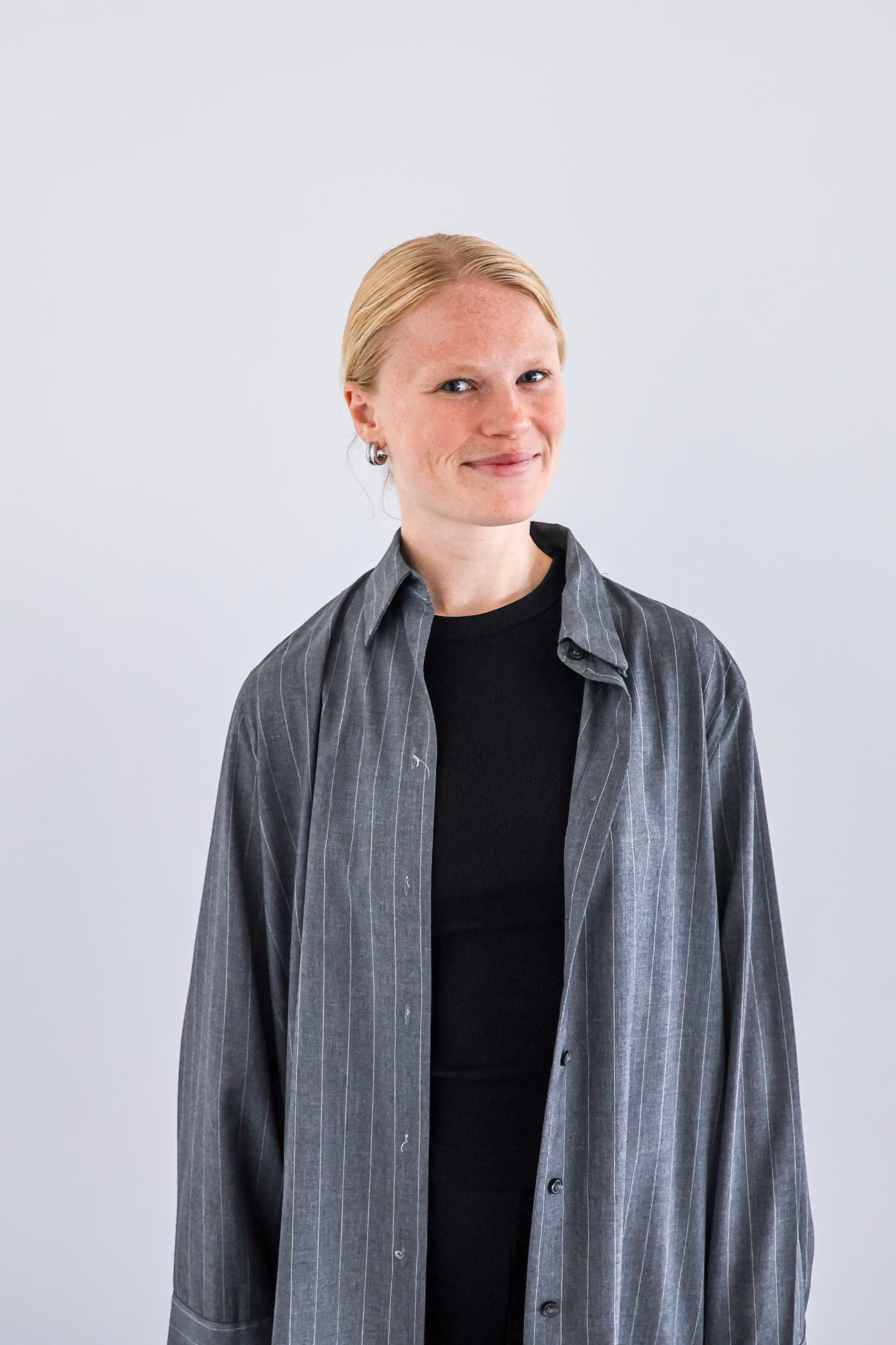 Lachende vrouw met blond haar in een lage knot draagt een donkergrijze, gestreepte, open overhemd over een zwarte top, met een lichtgrijze achtergrond.