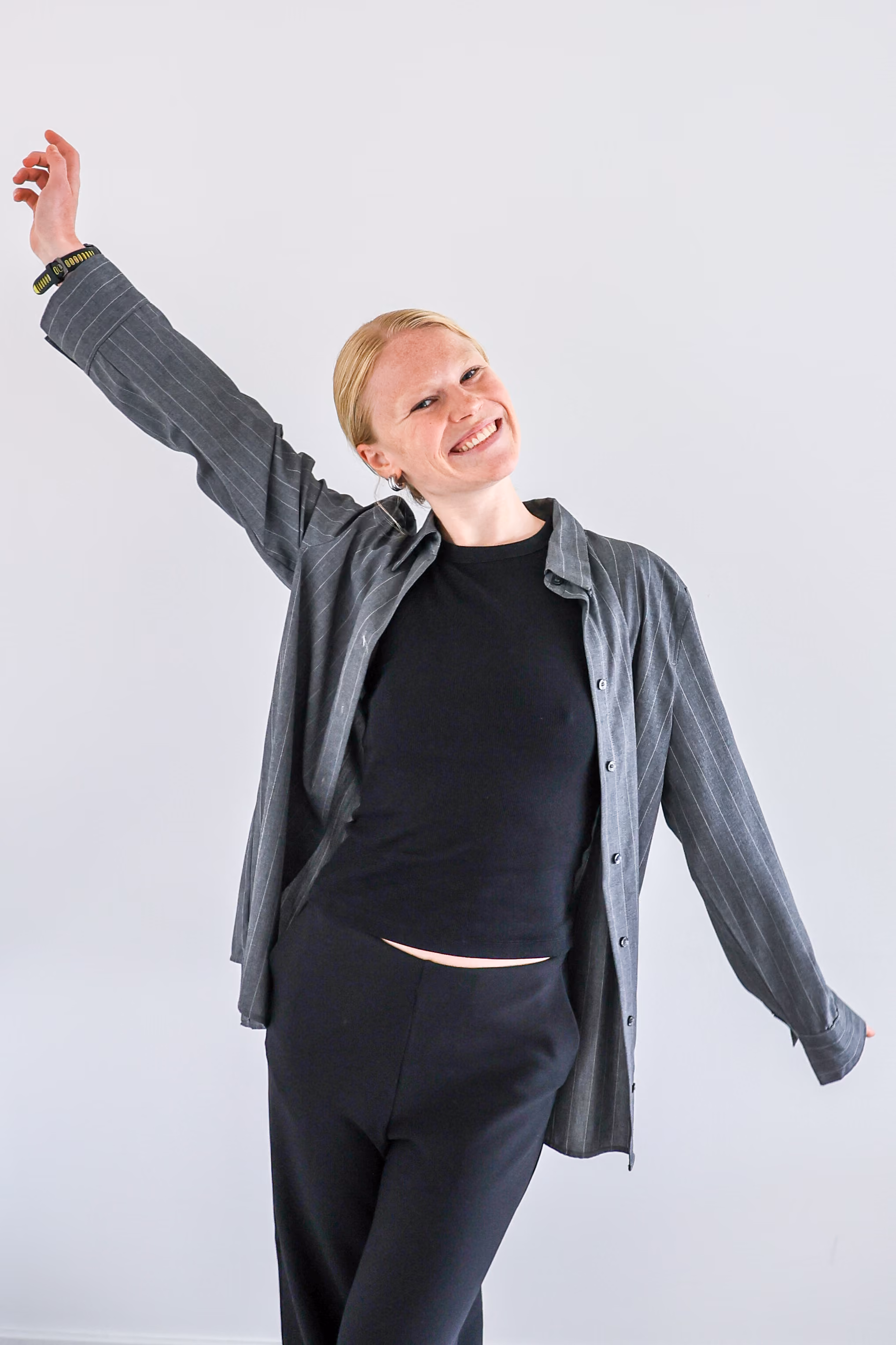 Vrouw die lacht en poseert met opgeheven armen, gekleed in een zwarte top, zwarte broek en een grijze gestreepte jas tegen een witte achtergrond.