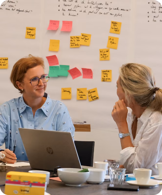 Twee vrouwen zitten aan een tafel in een kantoorruimte, ze praten met elkaar terwijl een laptop en koffiekopjes op de tafel staan. Op de achtergrond hangt een whiteboard met diverse kleurrijke sticky notes en handgeschreven aantekeningen.
