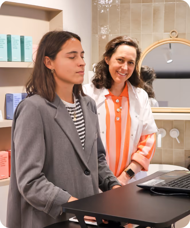 Twee vrouwen in een moderne winkel- of kantoorruimte, waarbij één vrouw met gesloten ogen voor een laptop aan een hoge tafel staat en de andere vrouw, die een witte jas draagt en een oranje-witte gestreepte trui onder heeft, glimlacht terwijl ze toekijkt.