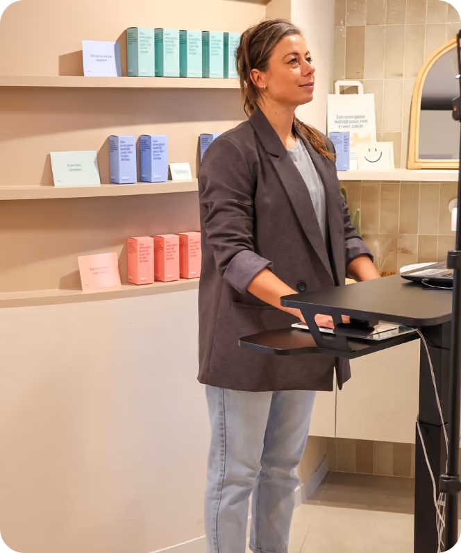 Vrouw staat bij een balie in een winkel met pastelgekleurde verpakkingen op planken achter haar.