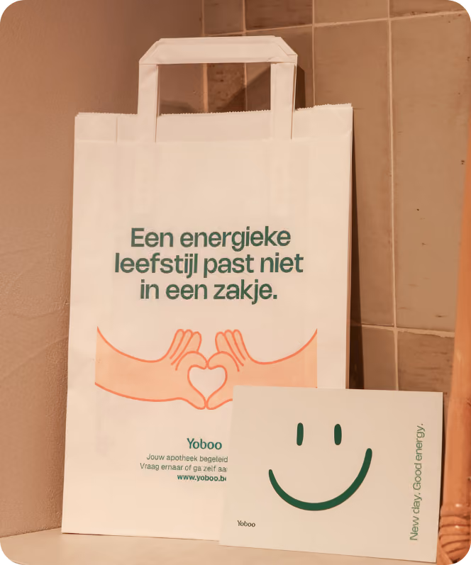 Een wit papieren zakje met groene tekst 'Een energieke leefstijl past niet in een zakje' en illustratie van twee handen die een hart vormen, naast een kaartje met een grote groene glimlach en tekst 'New day. Good energy.'