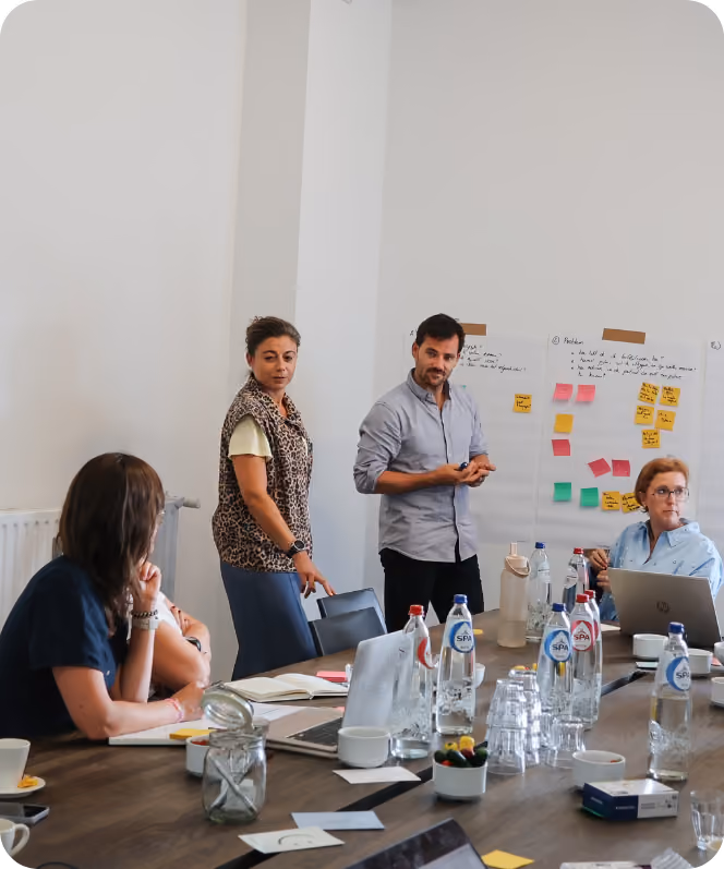 Vier volwassenen in een vergaderruimte met grote tafel waarop laptops, waterflessen, kopjes, glazen en notitieboekjes staan; twee personen staan bij een muur met grote vellen papier en kleurrijke plakbriefjes, terwijl de anderen aan tafel zitten en luisteren.
