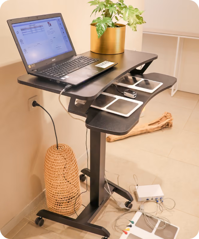 Een zwart verstelbaar bureau op wieltjes met een open Acer laptop, twee witte tablets en een USB-stick op het bureau, een gouden plantenpot met groene bladeren erop, en een gevlochten lampenkap ernaast tegen de muur.