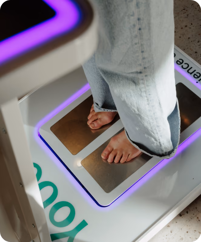 Iemand staat op blote voeten met een wijde spijkerbroek op een digitaal voetweegschaalapparaat met metalen platen en paars licht eromheen.