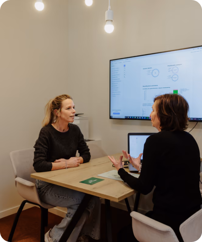 Twee vrouwen zitten aan een houten tafel in een kantoorruimte, in gesprek. Aan de muur hangt een groot scherm met grafieken en analysecijfers, en een laptop staat open op tafel.