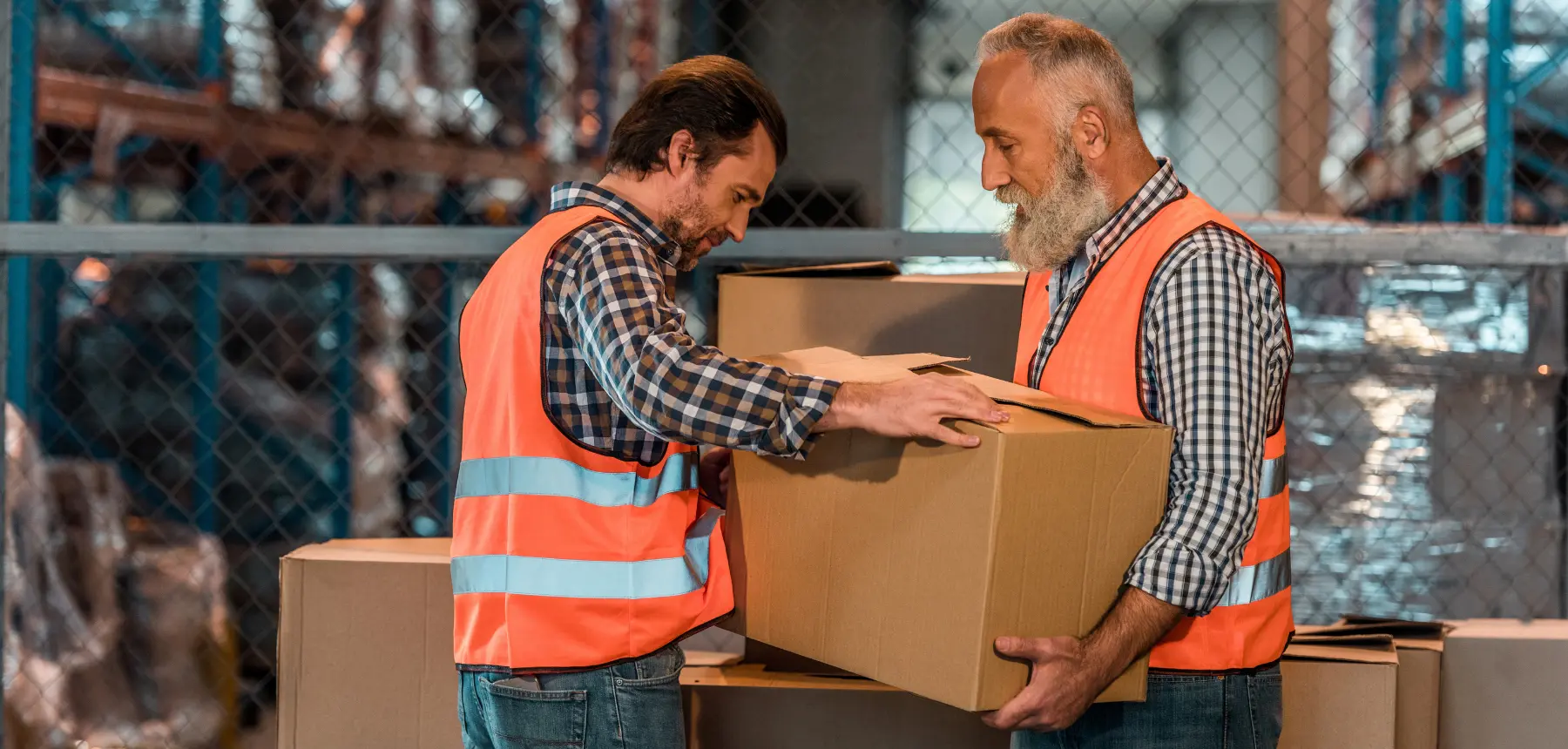 Logistics workers in safety vests handling a large cardboard box inside a warehouse, representing freight, shipping, and package handling operations.