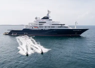 Large luxury yacht anchored at sea with two people riding jet skis in the foreground, leaving white trails on the water.