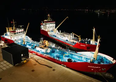 Nighttime aerial view of two red-and-white tankers moored side-by-side at a pier, blue deck lights glowing as cranes and hoses handle cargo on a dark harbor.