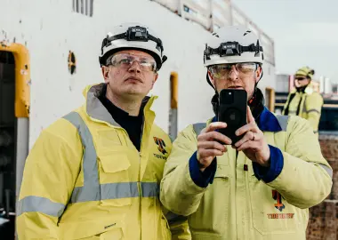 Two shipyard workers in yellow high-vis jackets, hard hats, and safety glasses stand on a vessel; one raises a smartphone to take a photo while the other looks on, with additional crew blurred in the background.