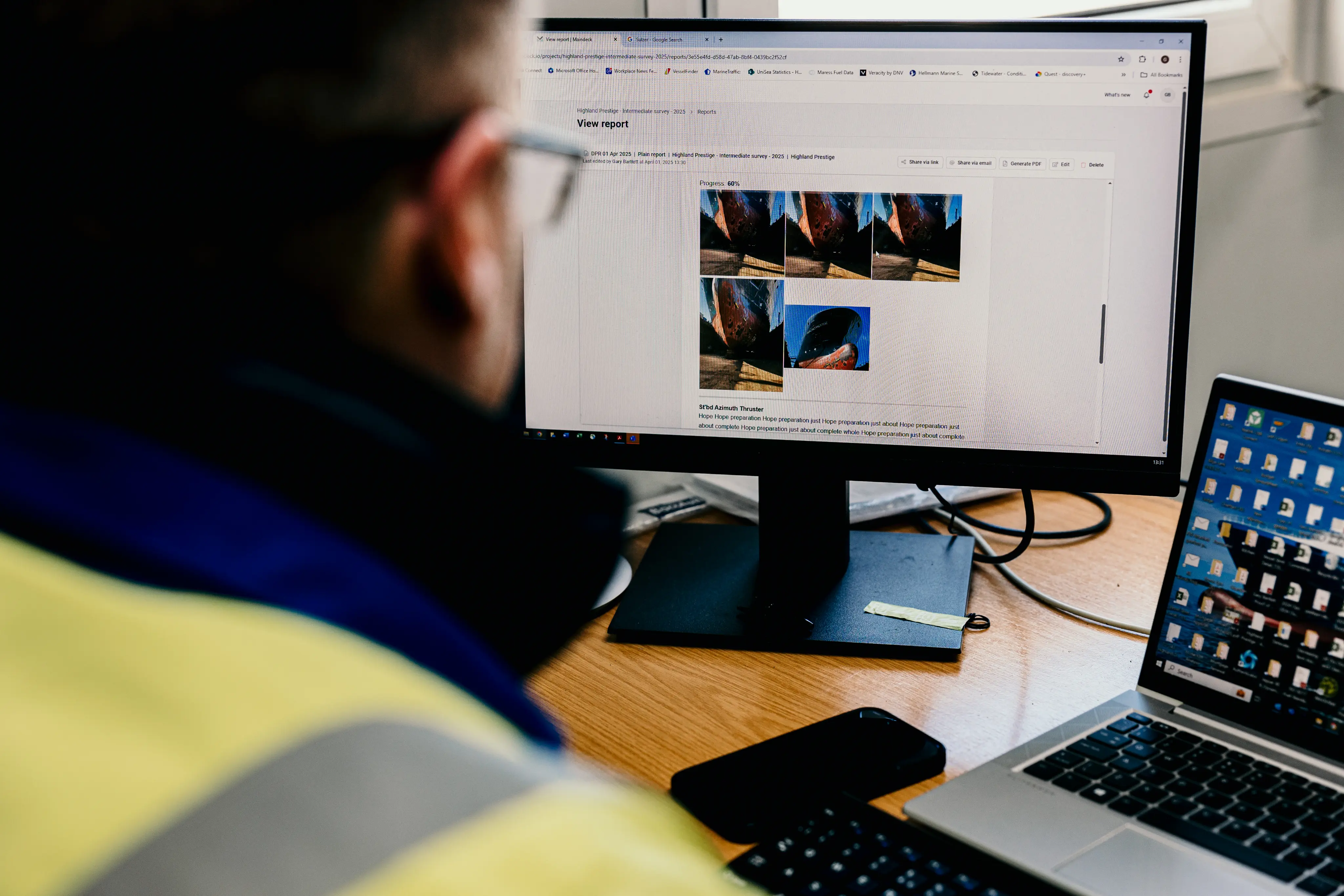 A worker in a high-vis jacket reviews a “View report” page on a desktop monitor showing multiple photos of a ship’s hull, with a laptop, keyboard, and phone on the wooden desk.