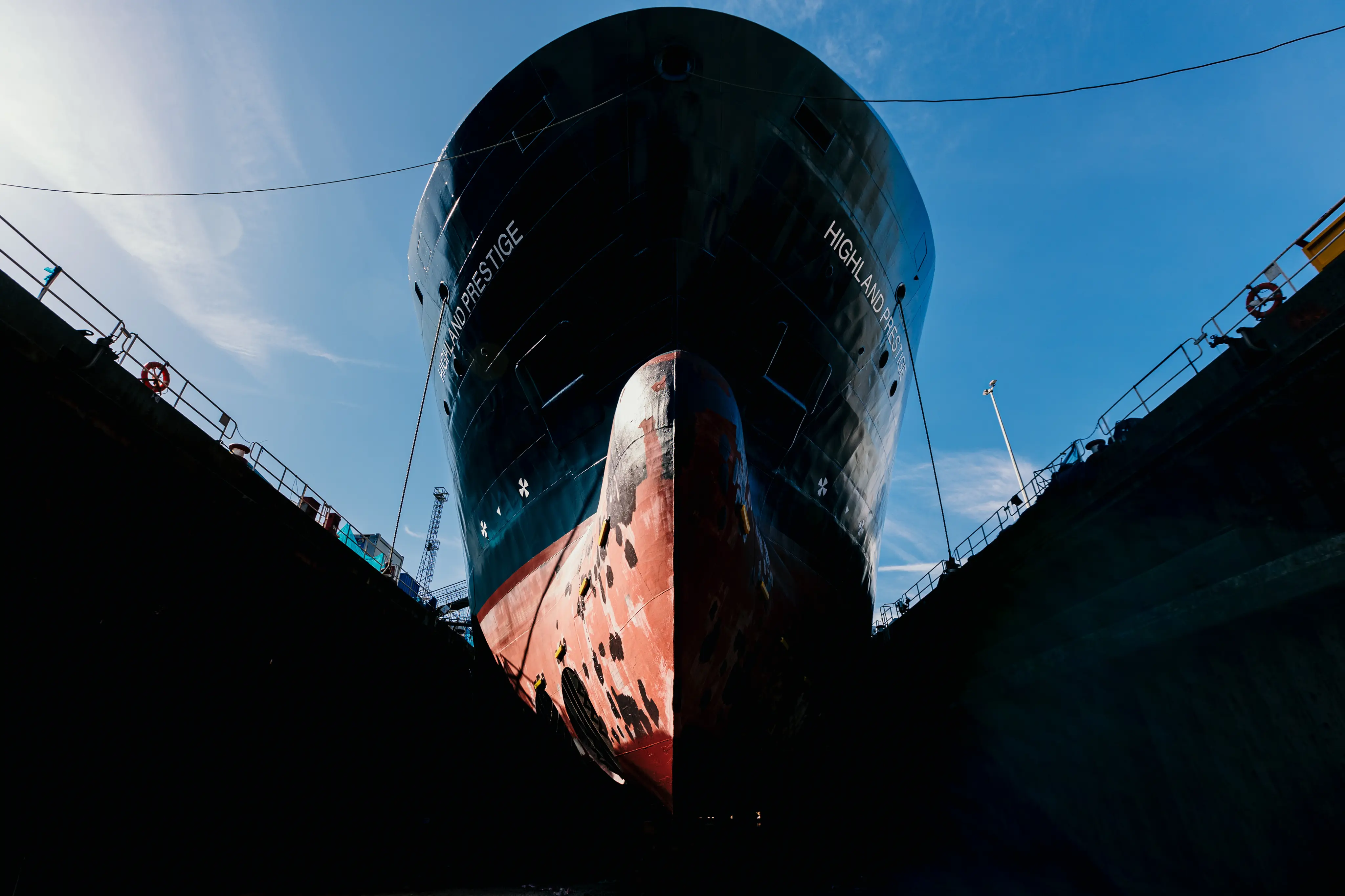 Bow-on view of the PSV Highland Prestige sitting in a dry dock, its dark hull and red, patchy antifouling exposed between high dock walls under a clear blue sky.