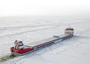 A red-and-white Wagenborg multipurpose cargo ship steams through a field of broken sea ice, seen from above, its long deck of hatch covers stretching between bow and stern.