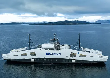 Side view of a white Norled ferry on dark blue water, with low islands and distant mountains beneath a cloudy sky.