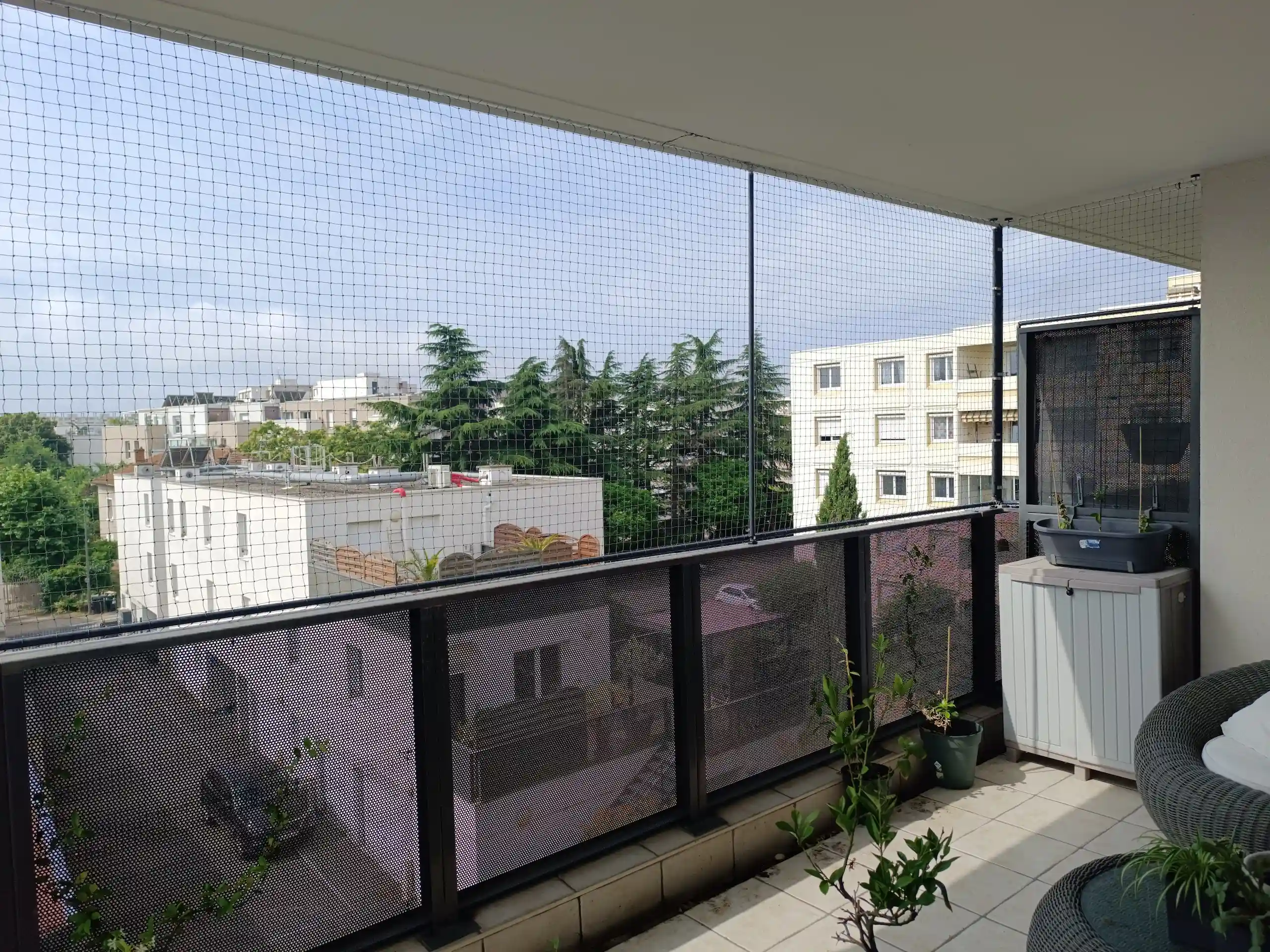 Balcon d'appartement avec filet de sécurité, plantes en pot, fauteuils en osier et vue sur des bâtiments résidentiels et des arbres.