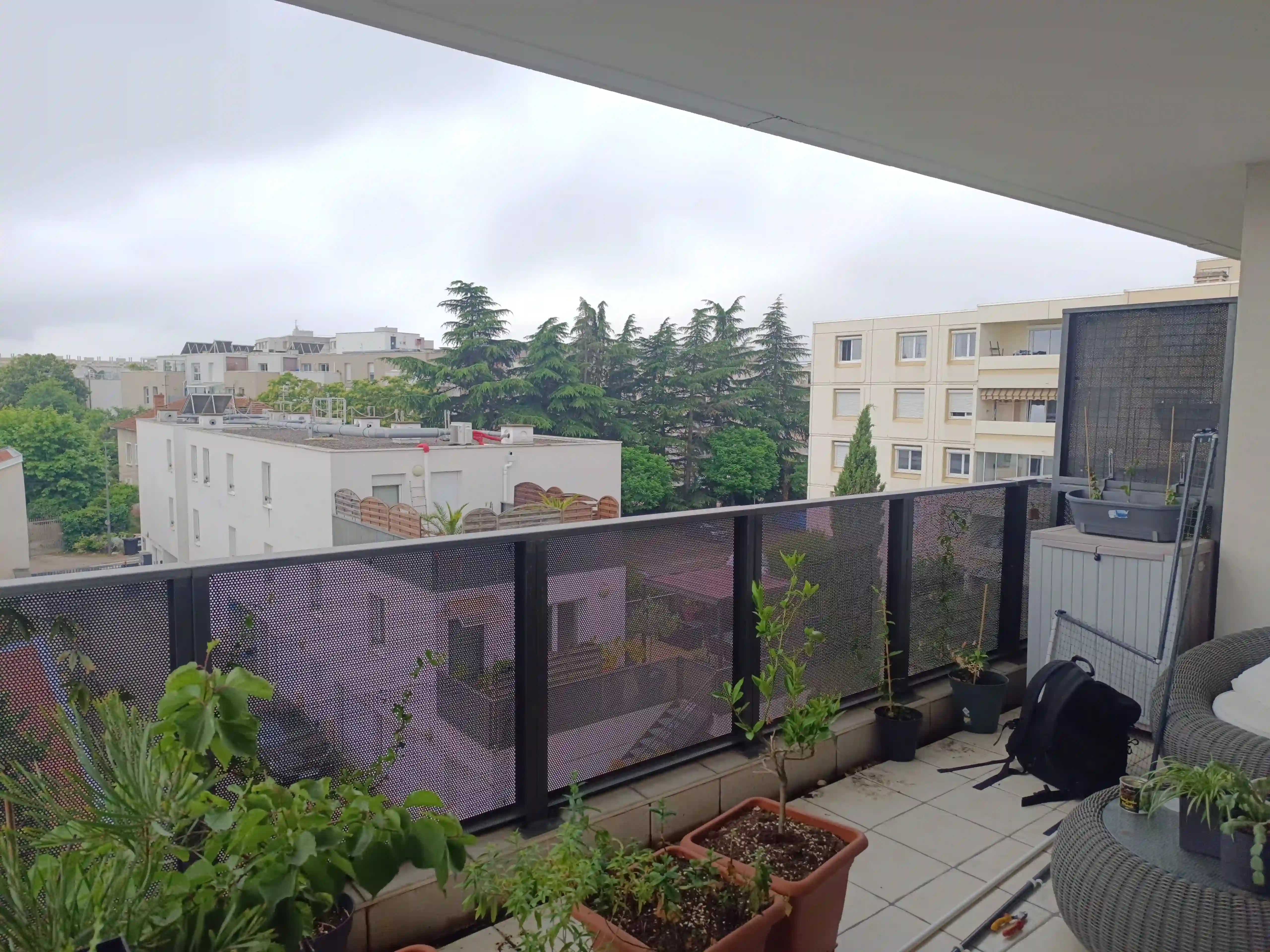 Balcon d'appartement avec des plantes en pots, un fauteuil en rotin, une table basse et une vue sur des bâtiments résidentiels et des arbres sous un ciel nuageux.