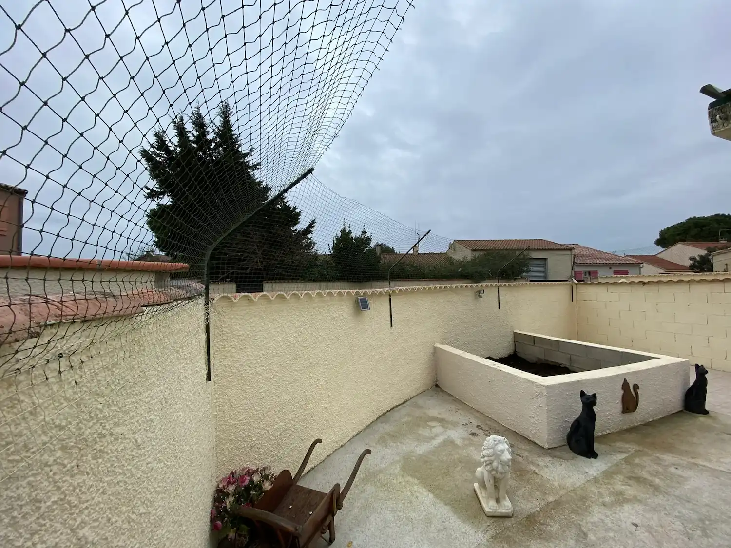Jardin clôturé avec un filet de protection au-dessus, statues d'un lion et de chats, et une brouette en bois.