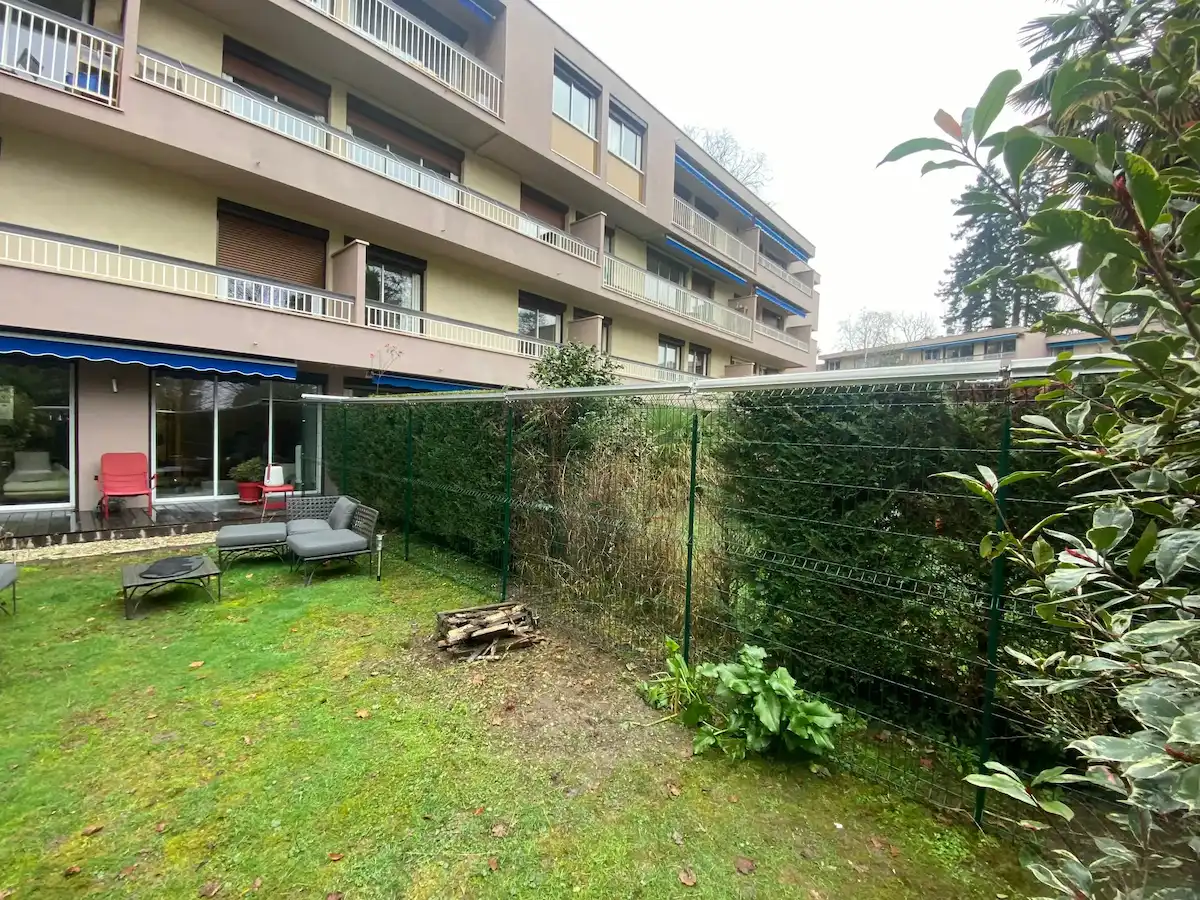 Jardin d'appartement avec pelouse verte, meubles d'extérieur gris, une pile de bois, une clôture métallique et des haies.