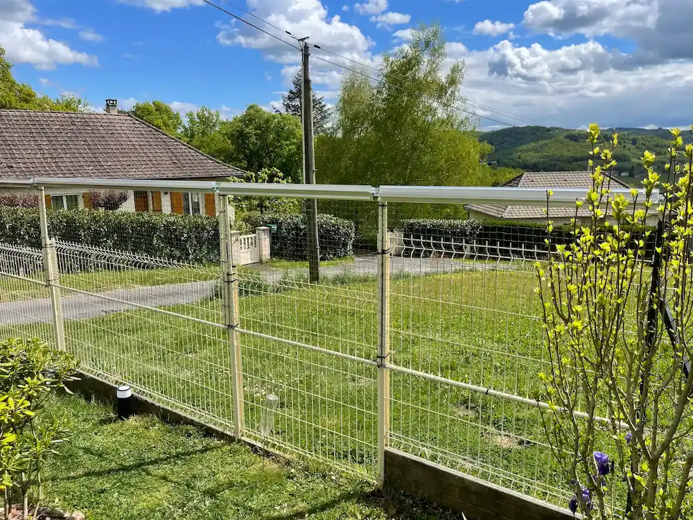 Clôture en métal blanc sur pelouse avec maison et arbres en arrière-plan sous un ciel bleu partiellement nuageux.