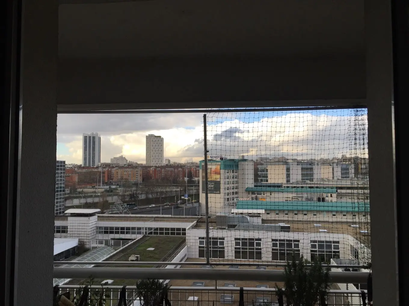 Vue urbaine depuis un balcon avec une balustrade et un filet de protection, montrant des bâtiments modernes sous un ciel nuageux.