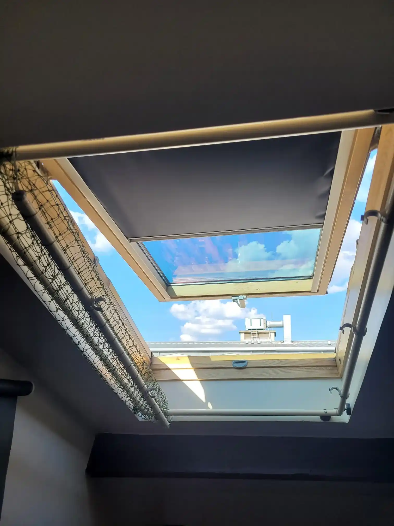 Fenêtre de toit ouverte avec un store partiellement déroulé laissant passer une vue sur un ciel bleu avec des nuages.