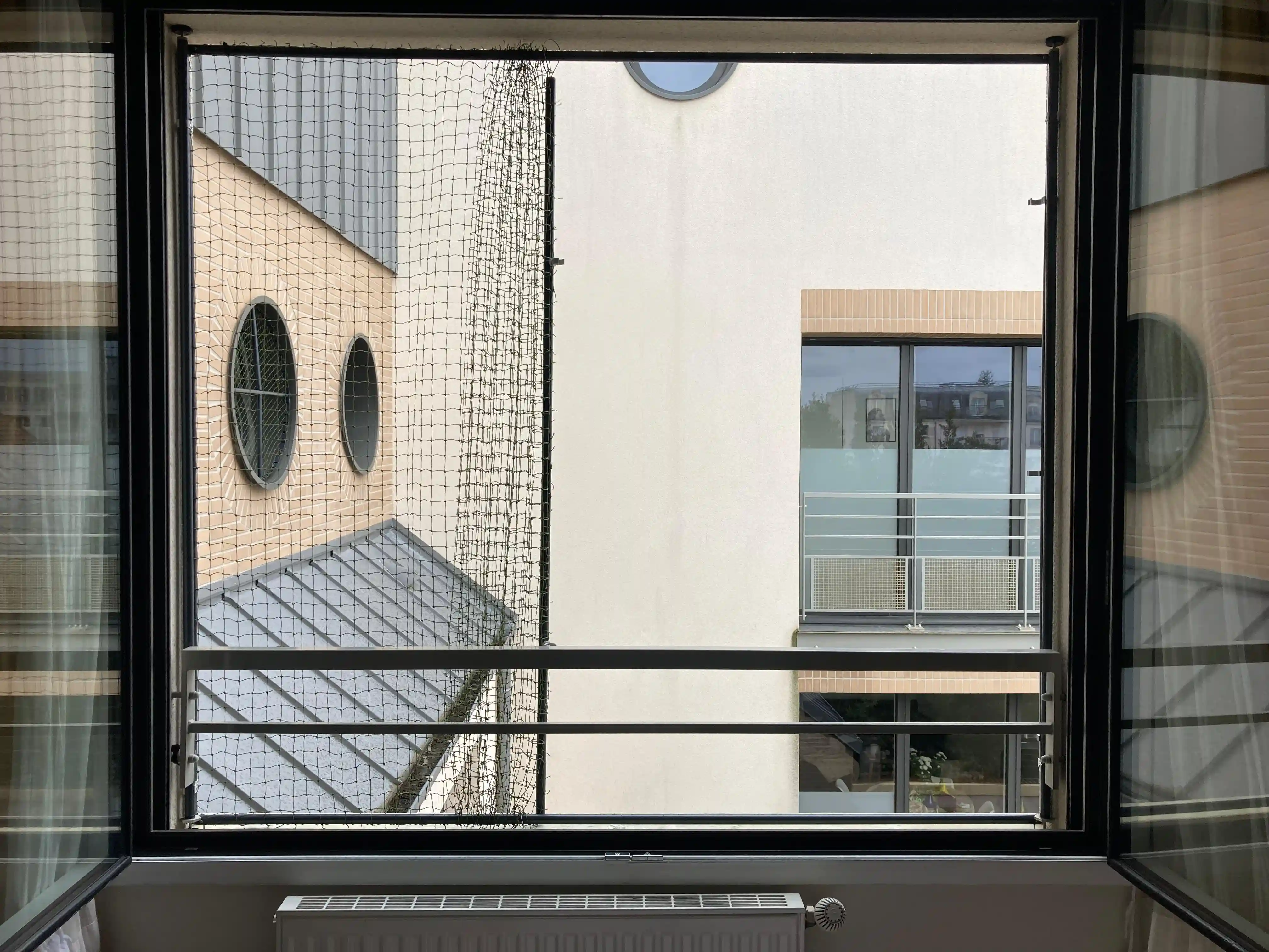 Fenêtre ouverte avec une barrière de sécurité en métal et un filet anti-chute, vue sur un bâtiment avec fenêtres ovales et rectangulaires.