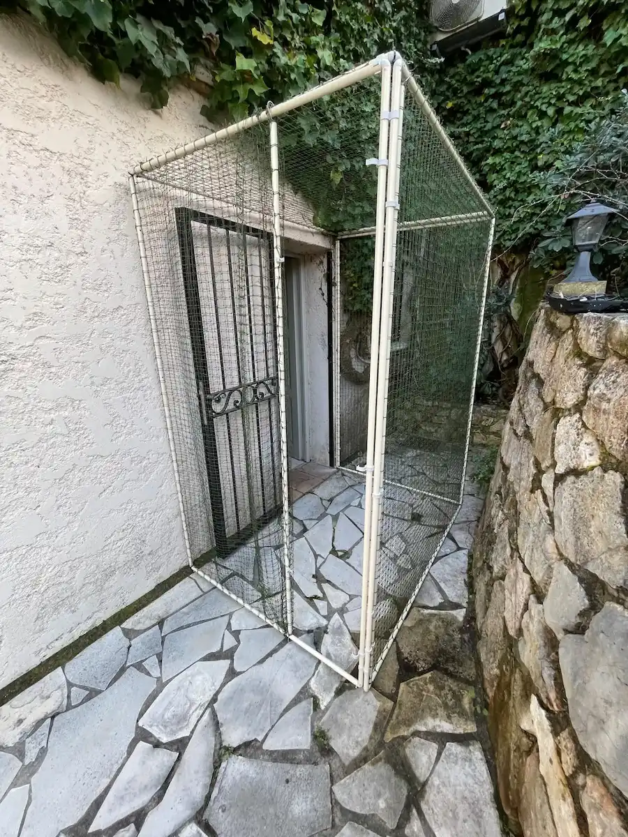 Structure en PVC grillagée encadrant une porte extérieure d'une maison avec un sol en pierres irrégulières et un mur en pierre à droite.