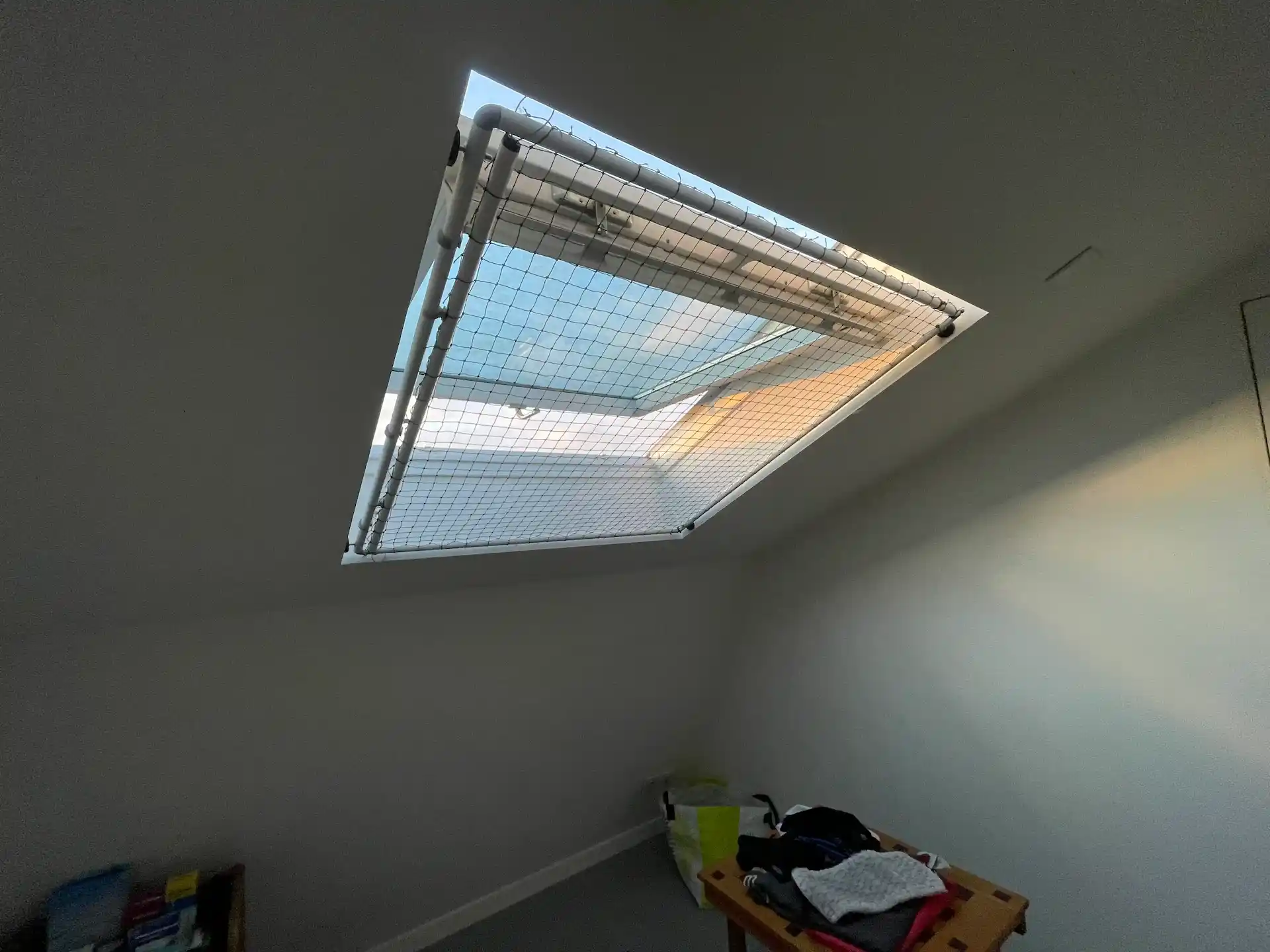 Une fenêtre de toit ouverte avec une moustiquaire sur un plafond incliné dans une pièce minimale avec une table et des vêtements pliés.