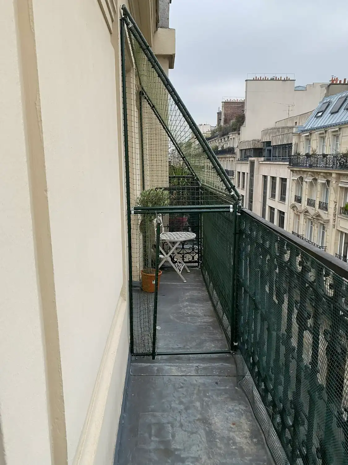Balcon étroit avec une table pliante blanche, une plante en pot, et un filet de protection vert entourant une partie du balcon.