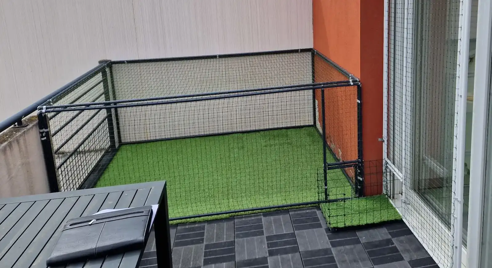 Pet balcony enclosed with black metal frame and netting on green artificial grass beside glass door.