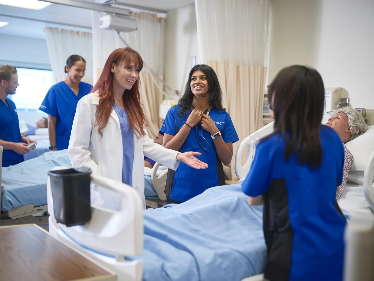A female doctor speaks to a group of medical students in blue scrubs gathered around a hospital bed with a patient, in a bright clinical setting.