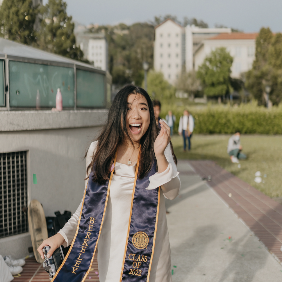 Lydia with a graduation sash, holding a camera, smiling.