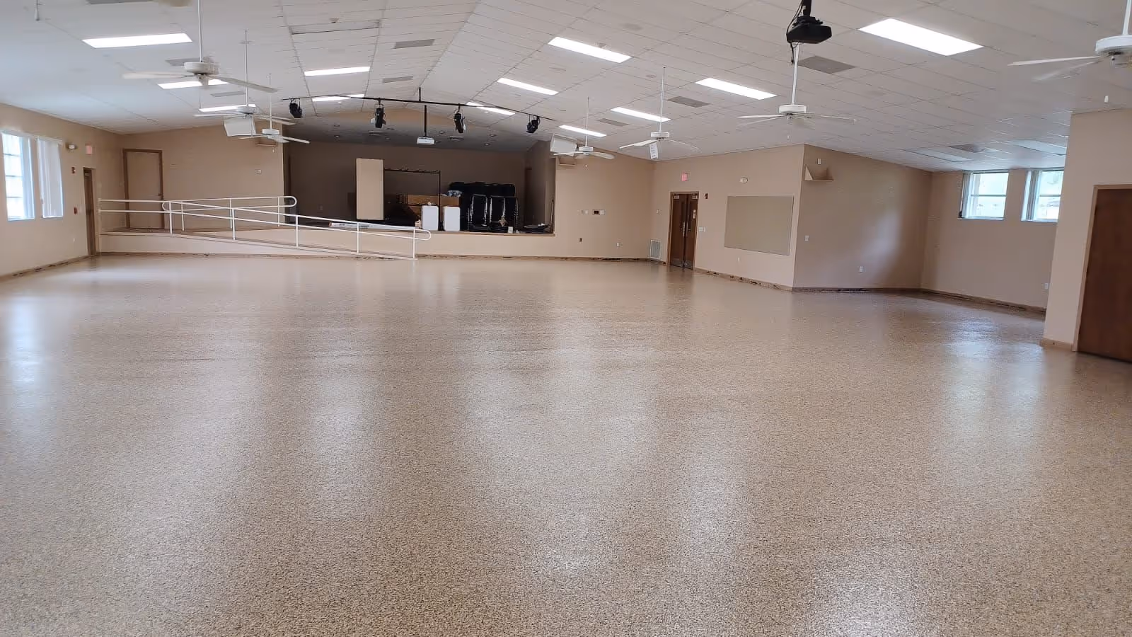 Empty large room with beige speckled flooring, ceiling fans, and a small stage with stacked chairs at the back.