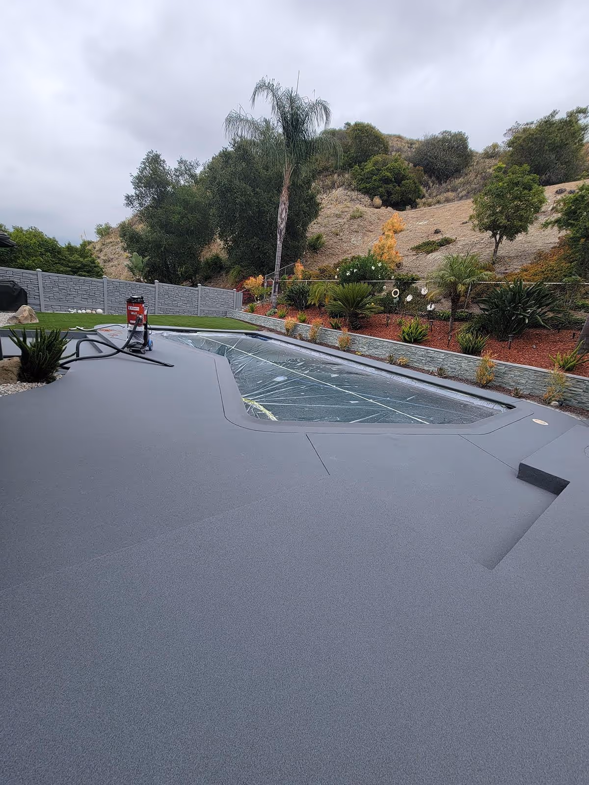 Outdoor swimming pool covered with a clear plastic safety cover surrounded by a smooth gray quartz pool deck, with landscaped hillside in the background under a cloudy sky.