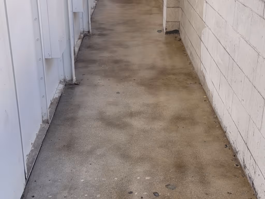 Narrow corridor with stained concrete floor flanked by a white wall with visible marks on one side and white paneling on the other.